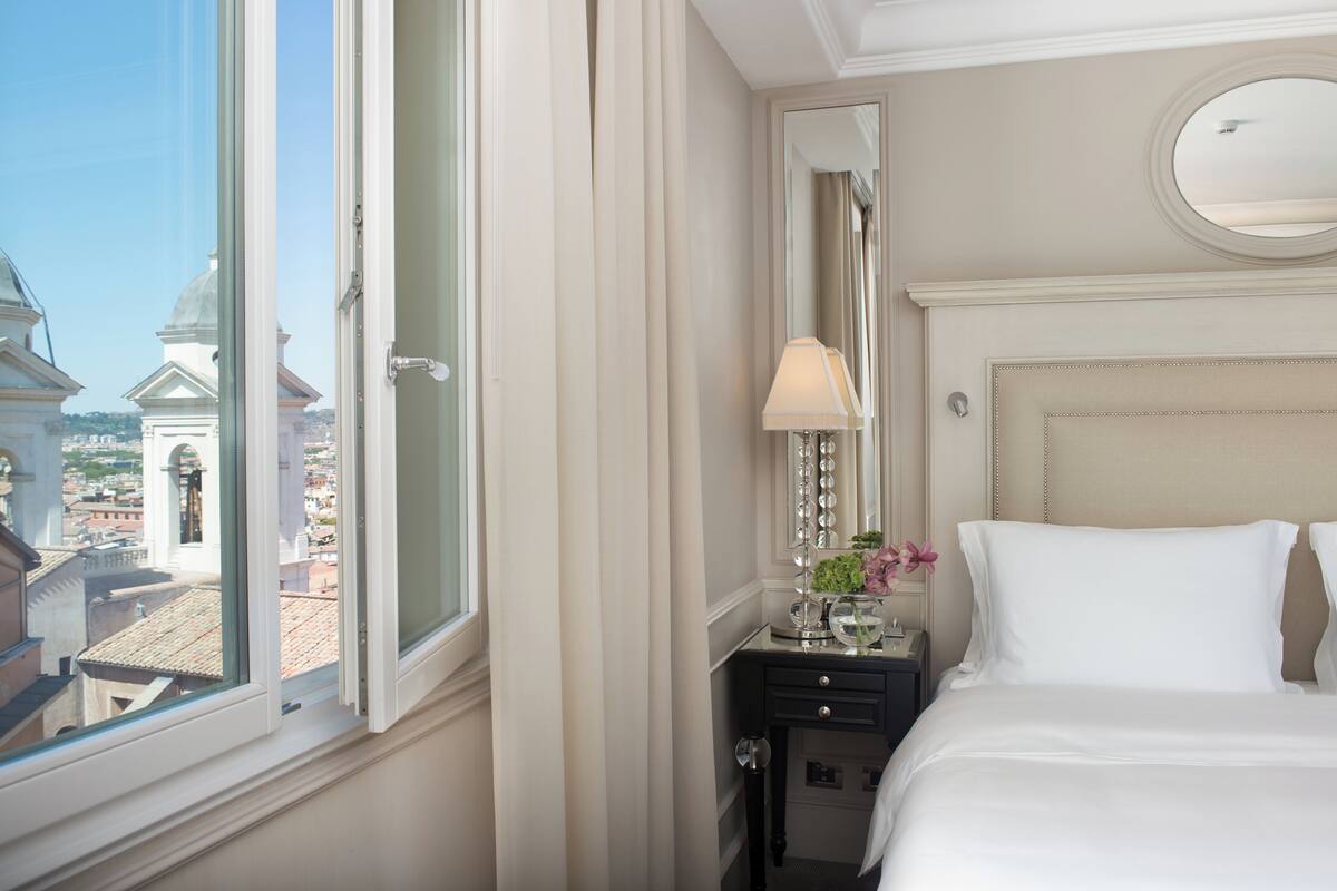 A hotel room with a large window, a bed, a nightstand, a lamp, and a mirror.