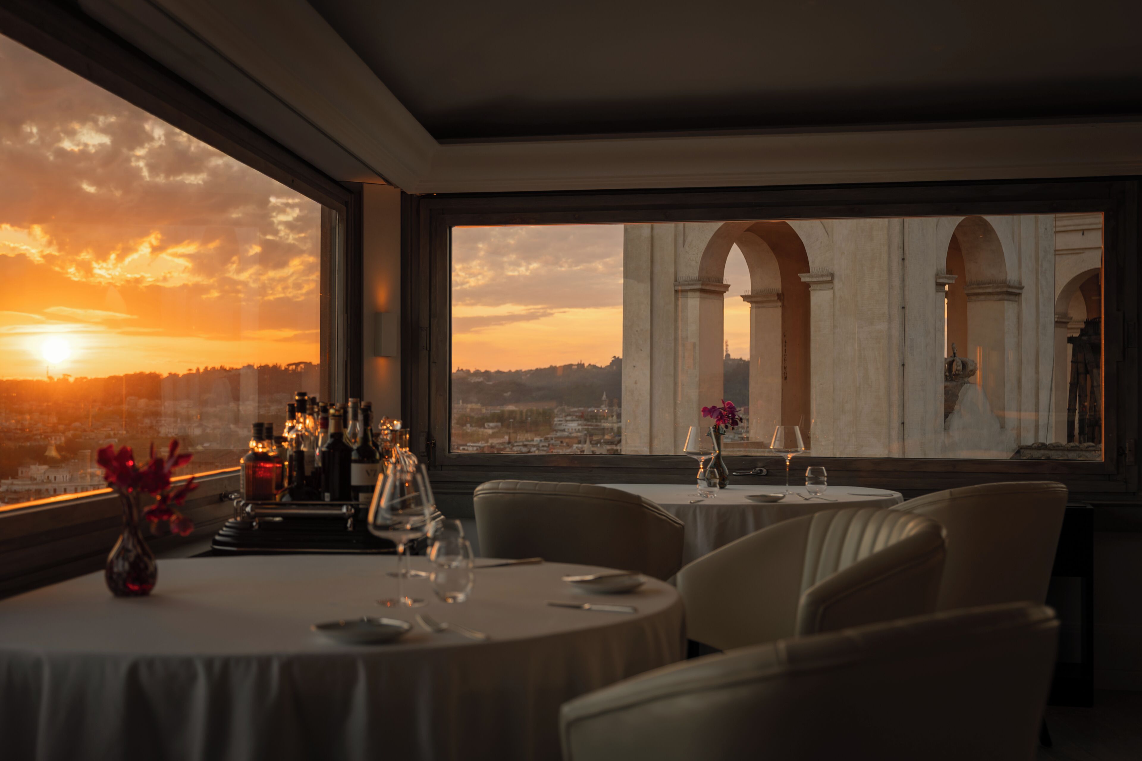 Un restaurante con mesas redondas, vista al atardecer y un paisaje urbano de fondo.