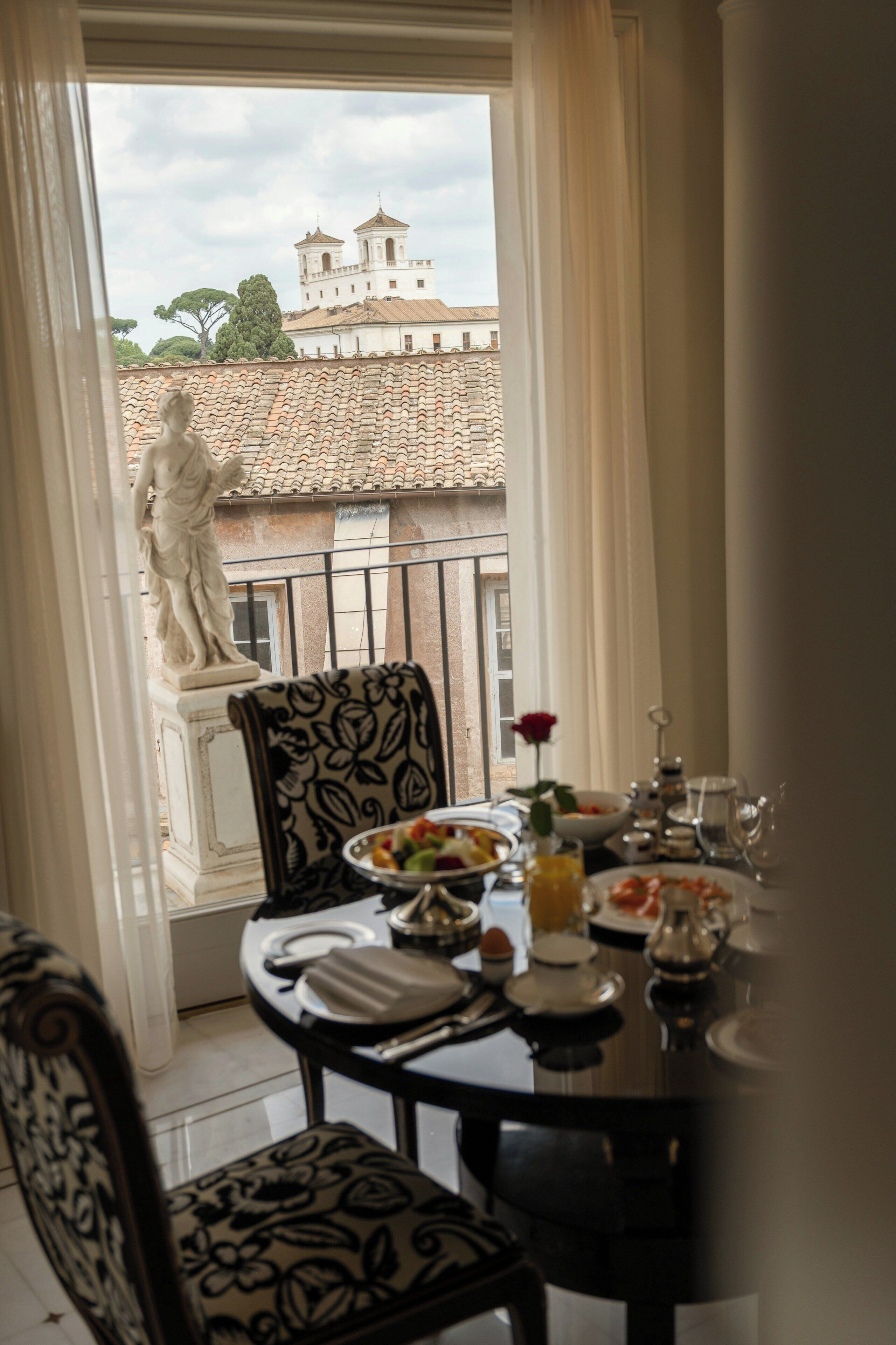Mesa de comedor con platos, vasos y una rosa en una habitación con vista a un edificio y una estatua.