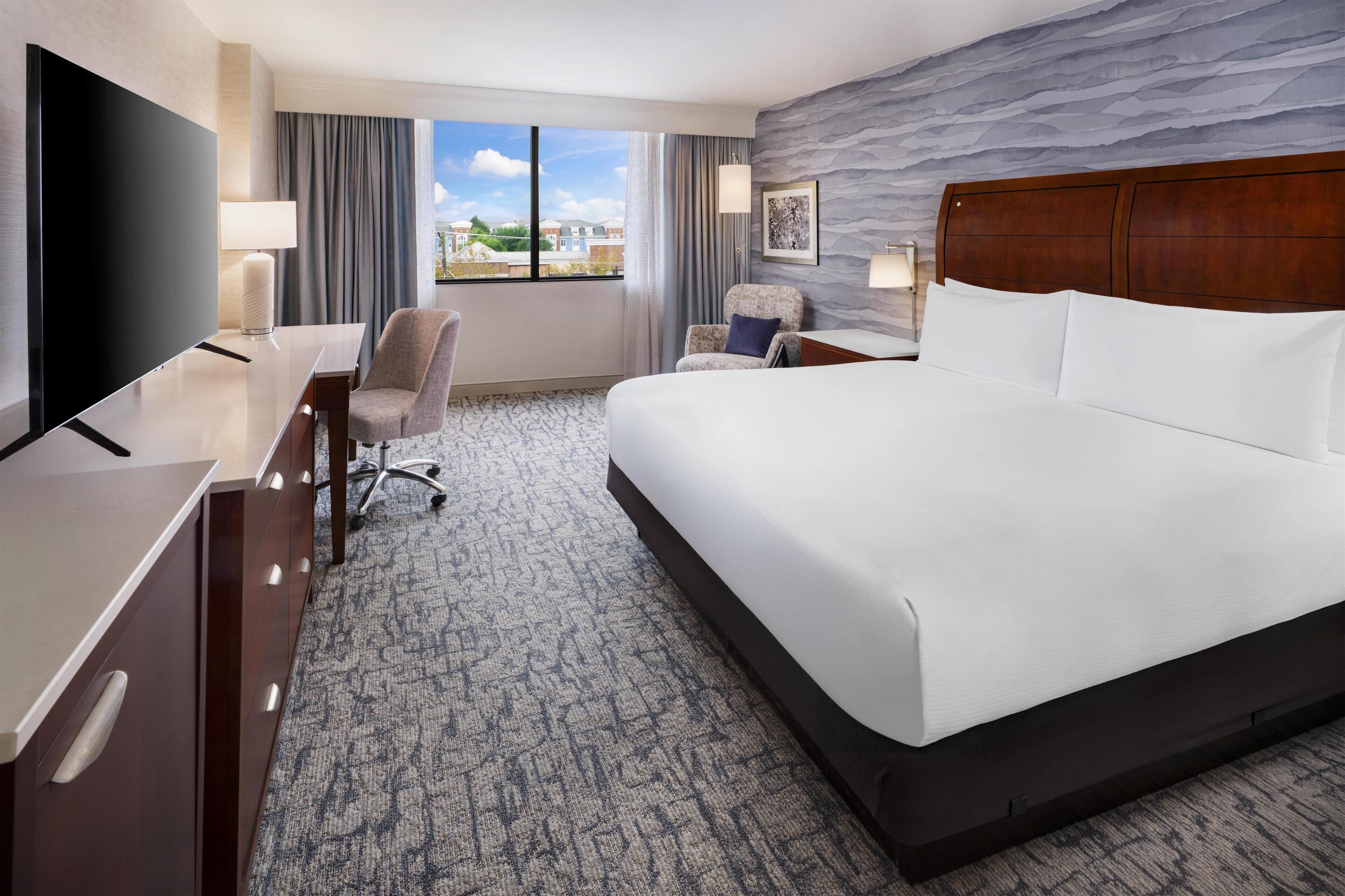 A hotel room with a large bed, a desk with a flat-screen TV, a chair, and a view of the outside.