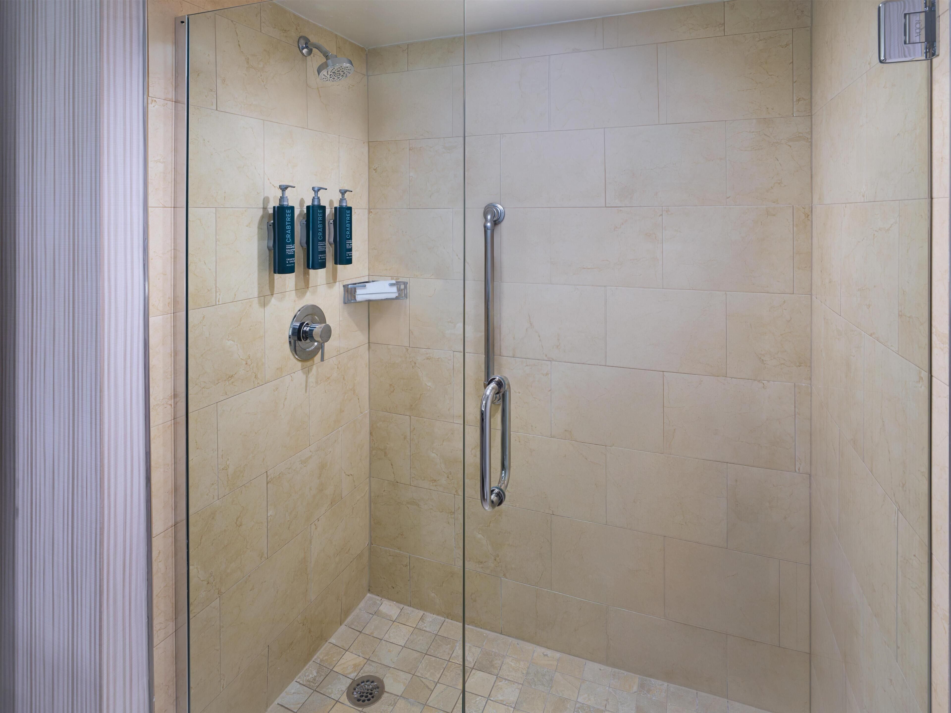 A walk-in shower with beige tiles, a glass door, a handheld showerhead, a handheld showerhead, and three bottles of shampoo.