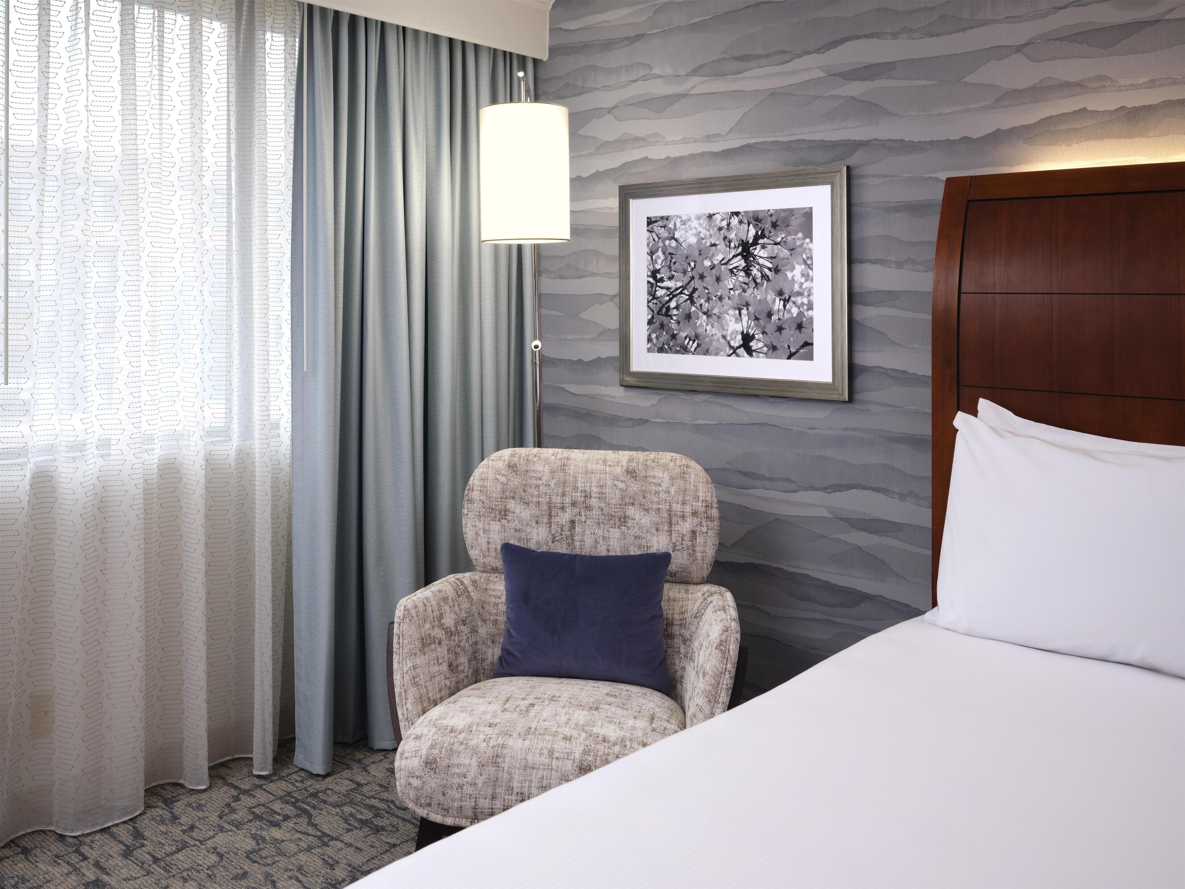Een hotelkamer met een bed, een stoel, een lamp en een lijst met een afbeelding aan de muur.