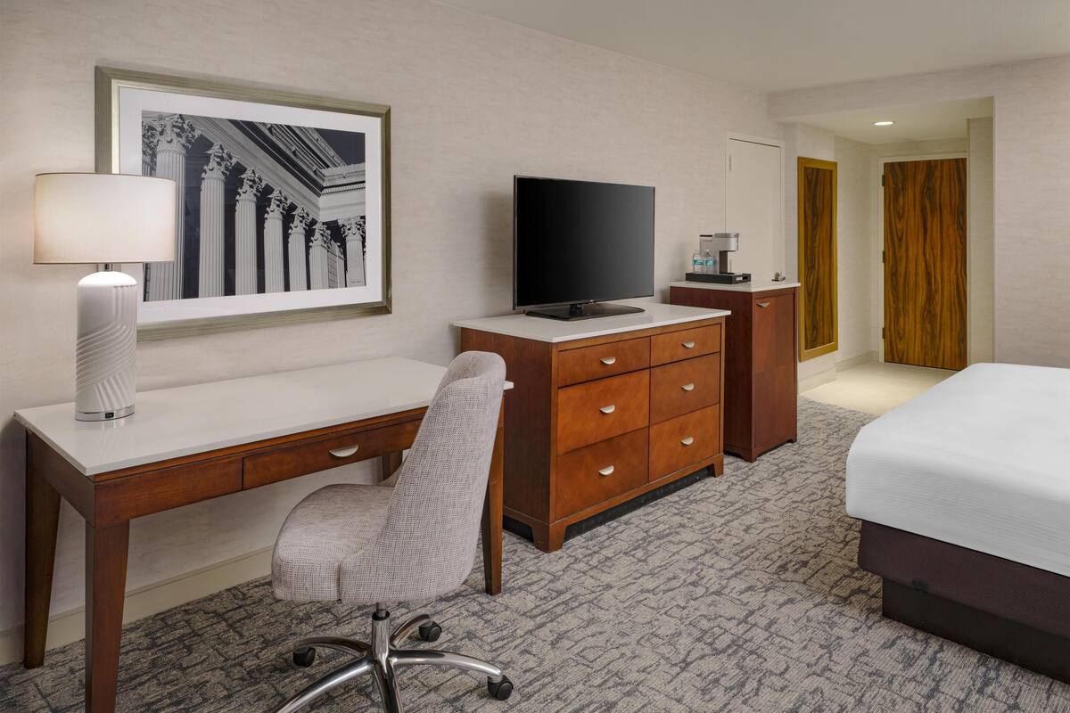 Una habitación de hotel con un escritorio de madera, una silla, un televisor, una cama y un cuadro en la pared.