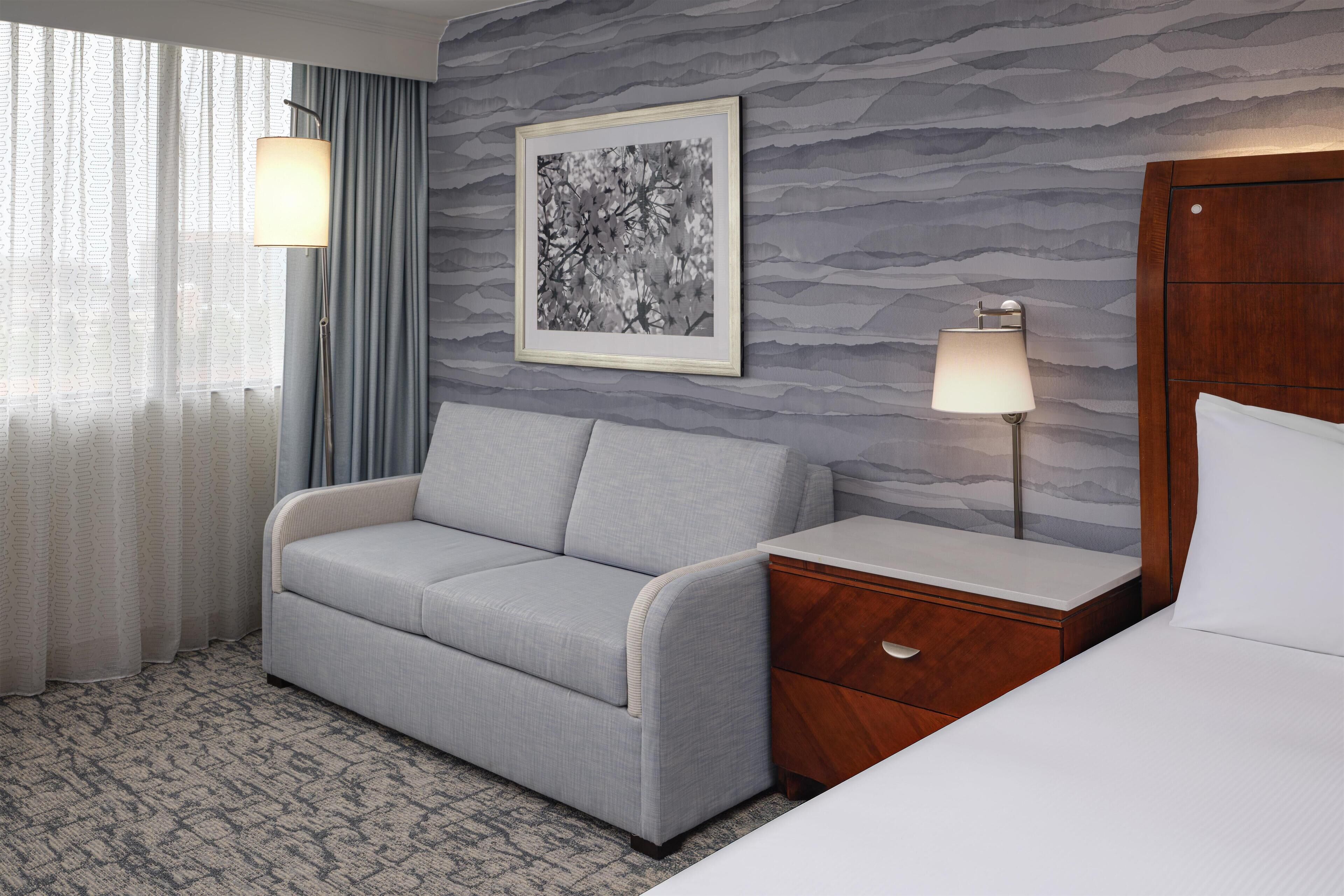 A hotel room with a grey sofa, a wooden nightstand, a framed picture on the wall, and a bed with white bedding.