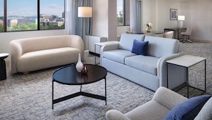 A hotel room with a sofa, a coffee table, a dining table with chairs, and a view of the city through large windows. - Hilton Washington DC/Rockville Hotel & Executive Meeting Ctr (Rockville)