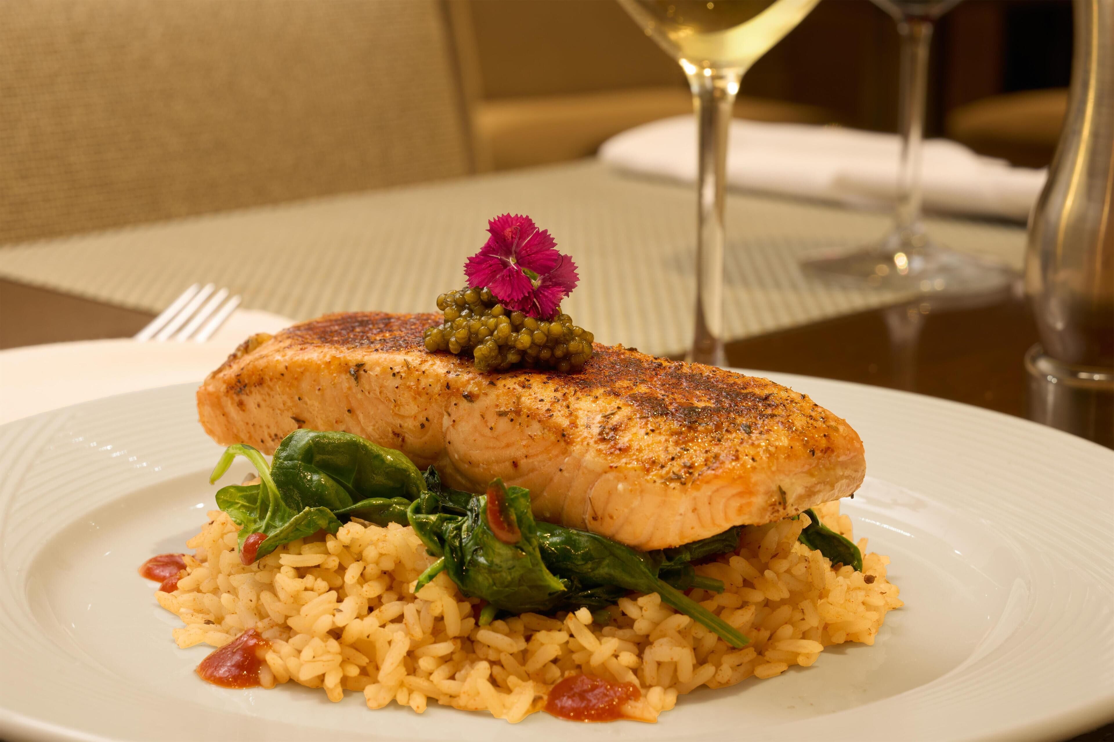 A plate of food with a piece of grilled fish, rice, and greens, accompanied by a glass of white wine.