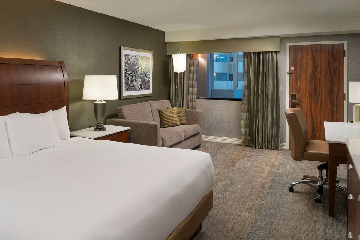 Habitación de hotel con una cama grande, un escritorio, una silla, una lámpara, un sofá y una ventana con cortinas.