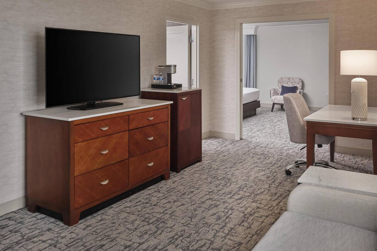 Habitación de hotel con televisor de pantalla plana, cómoda de madera, escritorio con lámpara, silla y cama con mesita de noche.