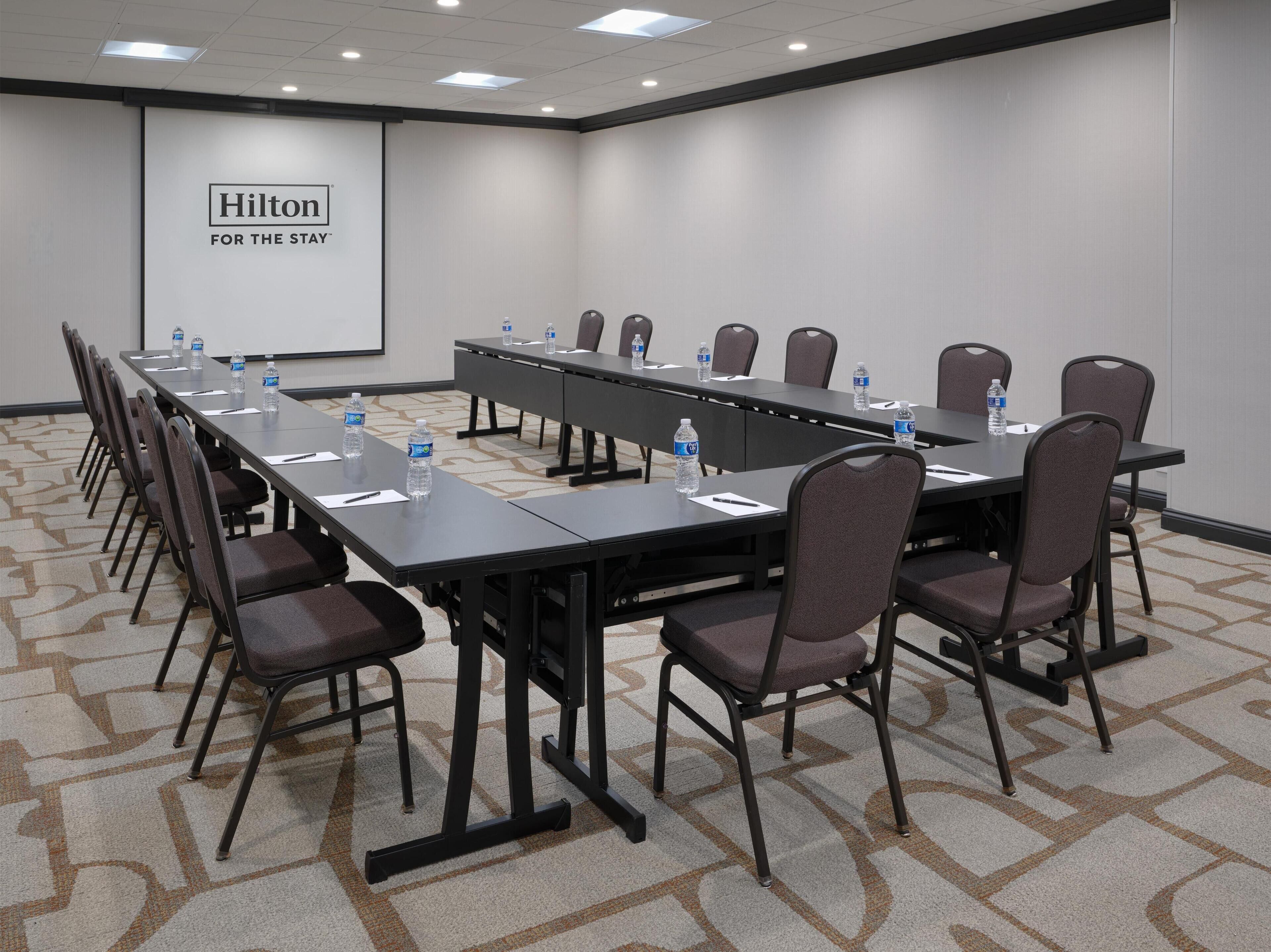 Uma sala de conferências com uma mesa comprida, cadeiras, garrafas de água e uma tela de projetor exibindo 'Hilton For The State'.