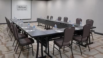 Uma sala de conferências com uma mesa comprida, cadeiras, garrafas de água e uma tela de projetor exibindo 'Hilton For The State'.