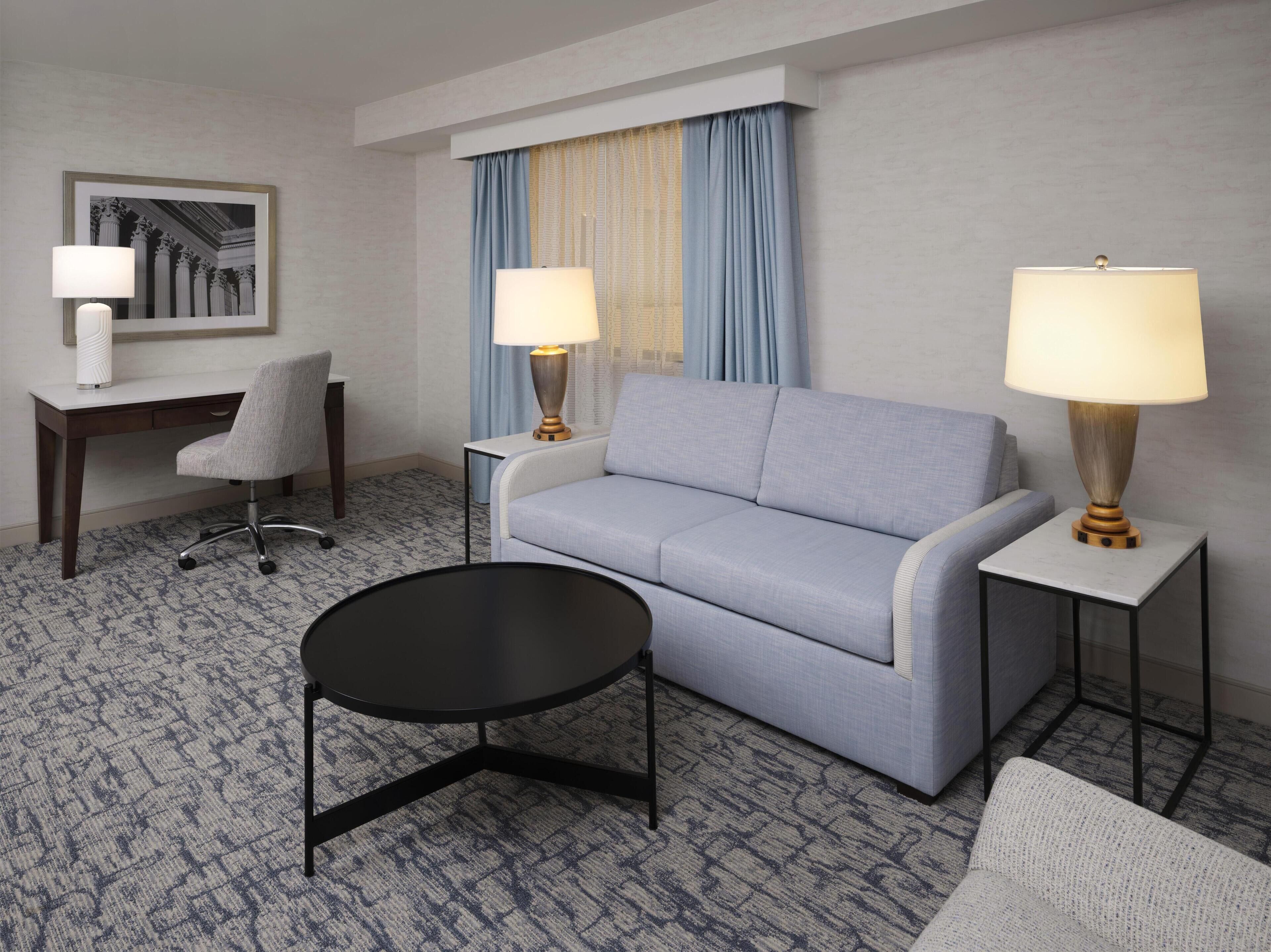Une chambre d’hôtel avec un canapé gris, deux tables d’appoint avec des lampes, une petite table ronde, un bureau avec une chaise et un tableau encadré au mur.