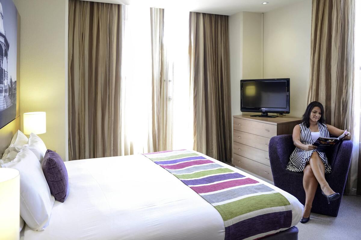 Una habitación de hotel con cama, televisor, sillón y cortinas.