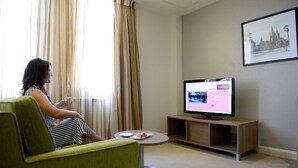 A woman sitting on a green armchair in a room with a television on a wooden stand, a small table with a plate of food, and a framed picture on the wall.