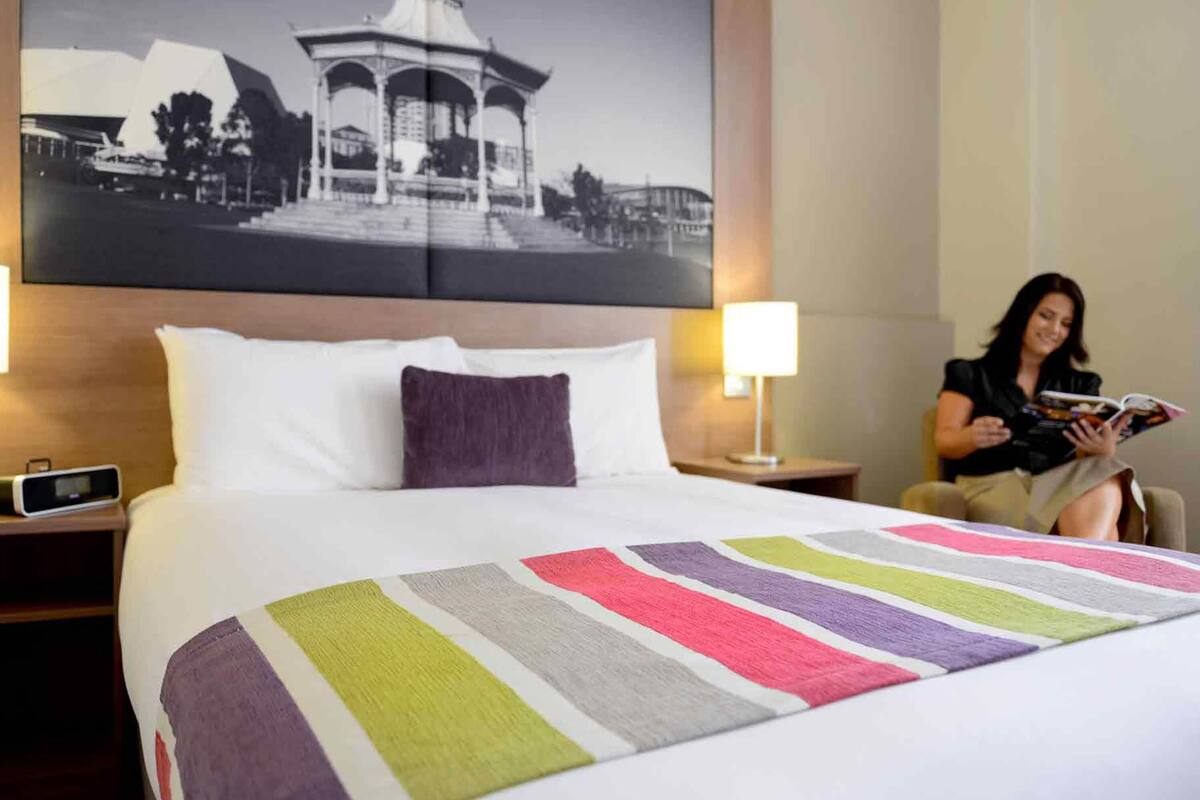 Habitación de hotel con cama, mesita de noche, lámpara y una persona leyendo una revista.