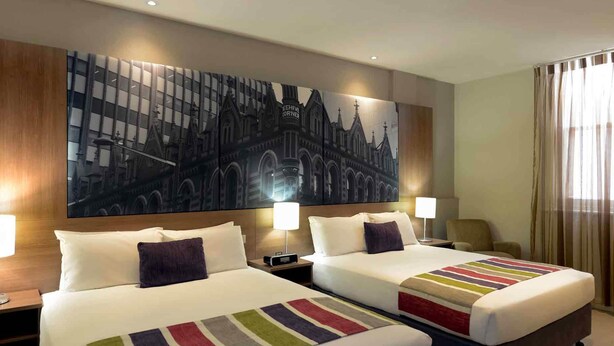 A hotel room with two beds, a large cityscape painting on the wall, and a view of the city through the window.