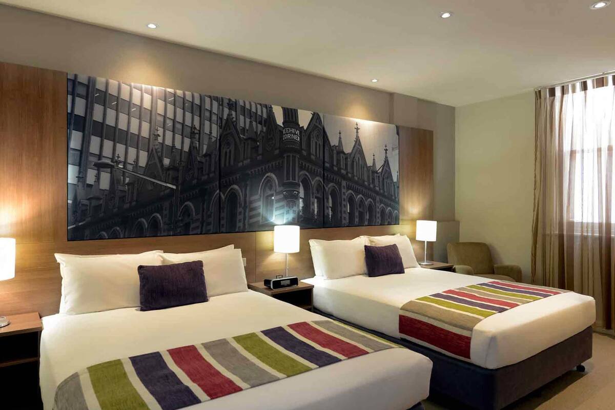 Habitación de hotel con dos camas, un cuadro grande con un paisaje urbano en la pared y vistas de la ciudad desde la ventana.
