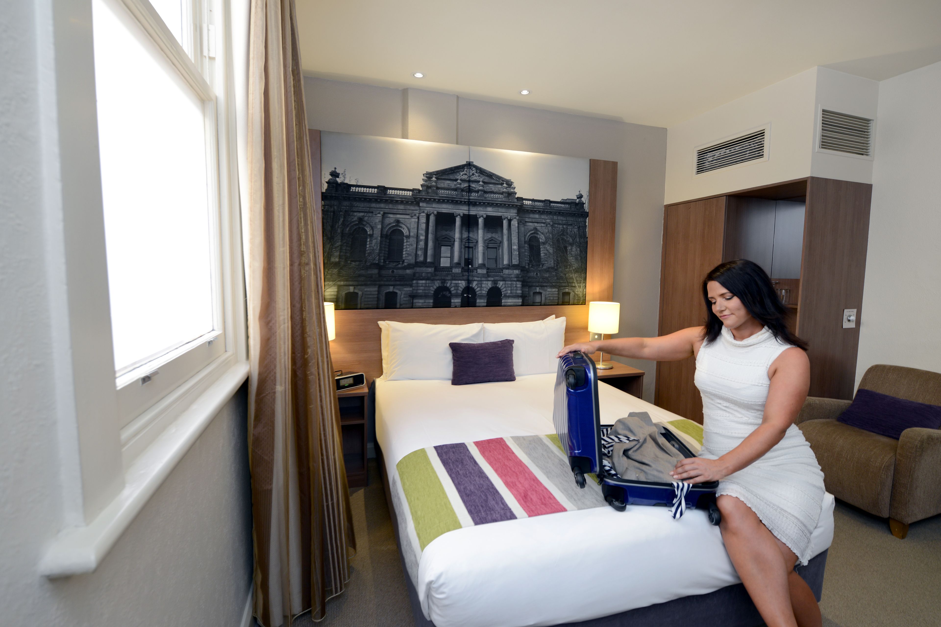 A hotel room with a bed, a large framed picture on the wall, a window, and a nightstand with a lamp.