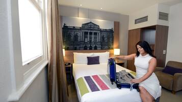 A hotel room with a bed, a large framed picture on the wall, a window, and a nightstand with a lamp.