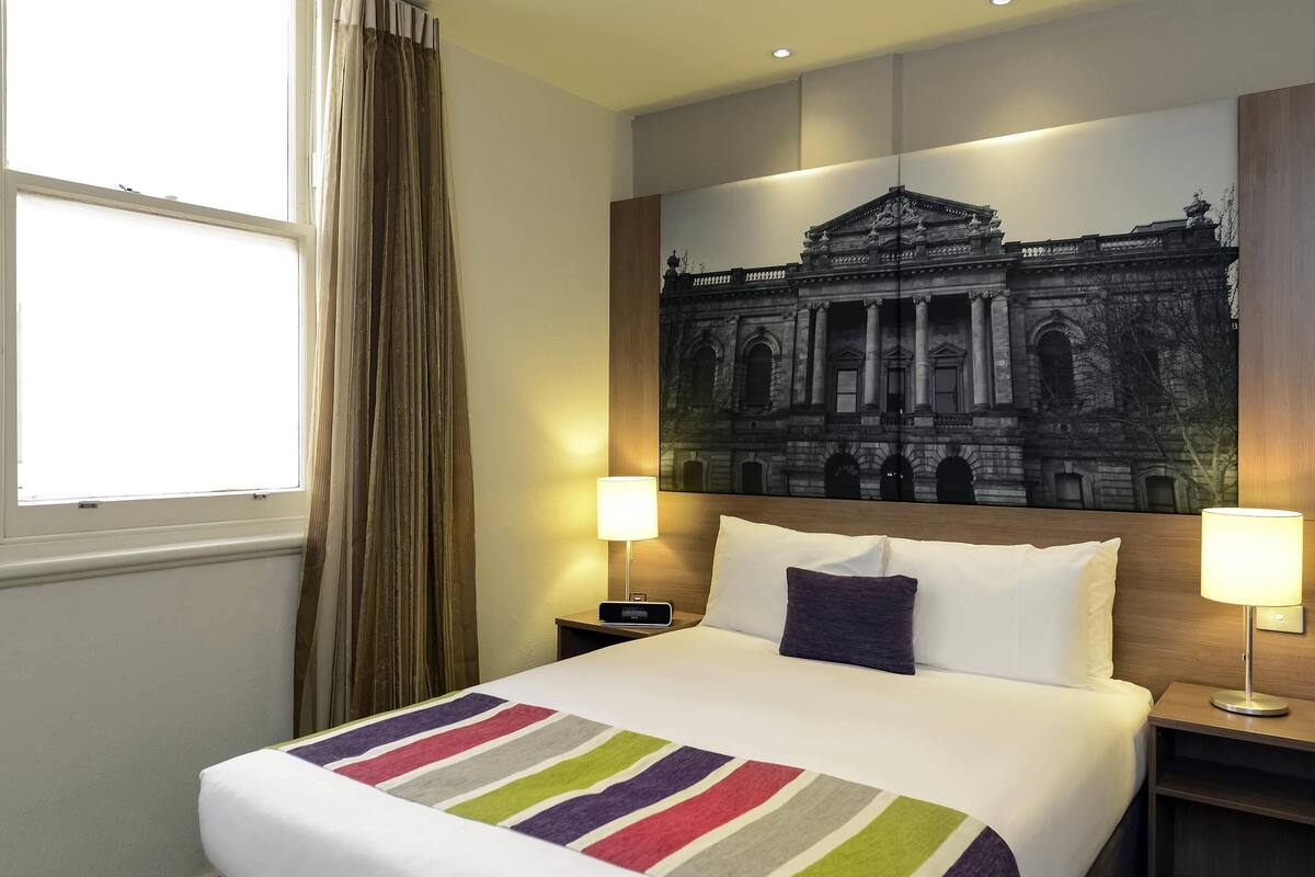 Habitación de hotel con una cama grande, dos lámparas de noche, una ventana con cortinas y una obra de arte en la pared que representa un edificio.