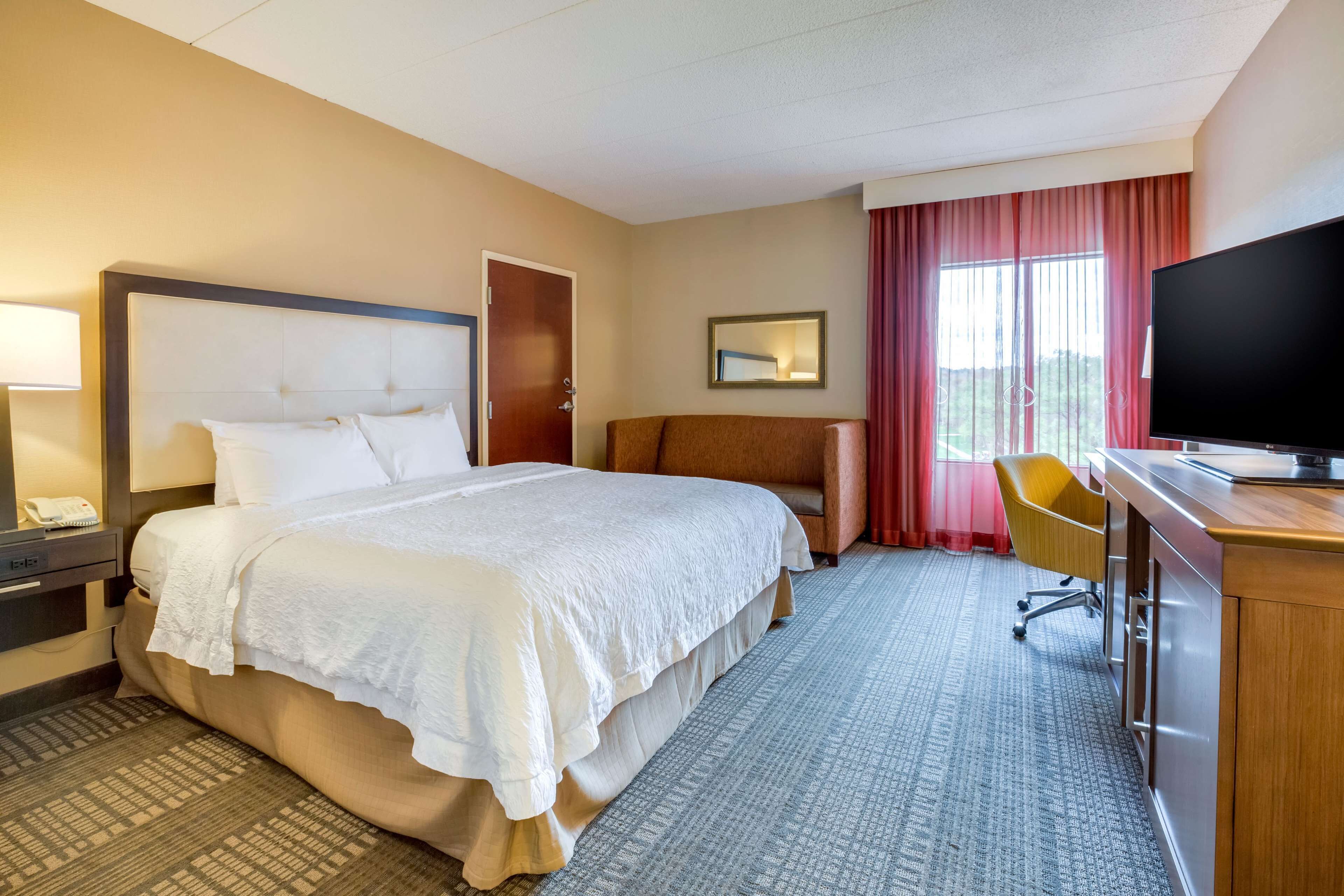 Quarto de hotel com uma cama grande, uma mesa de trabalho com televisão, uma cadeira e uma janela com cortinas.