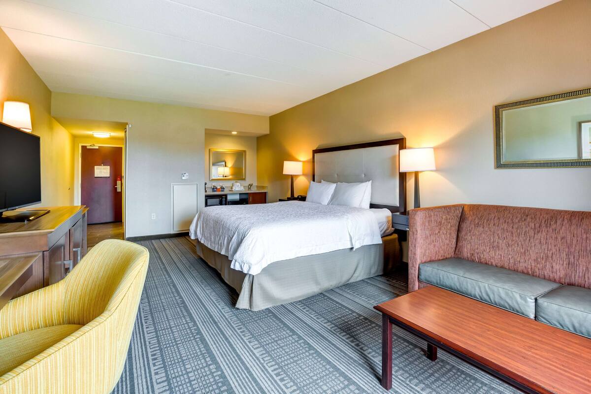 A hotel room with a bed, a sofa, a desk with a TV, a mirror, and a bathroom visible in the background.