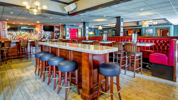 A bar area with a long counter, multiple stools, and a red booth seating area. - Hampton Inn Boston - Norwood (Norwood)