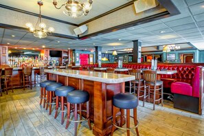 Un espace bar doté d’un long comptoir, de nombreux tabourets et d’un coin salon avec des banquettes rouges.