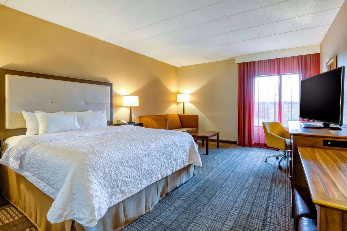 A hotel room with a large bed, two bedside lamps, a desk with a TV, a chair, and a window with curtains.