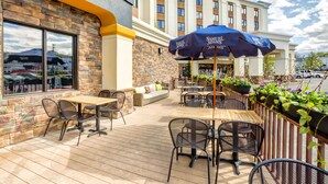 An outdoor patio area with wooden tables and chairs, a blue umbrella, and a view of the Hampton Inn hotel in the background. - Hampton Inn Boston - Norwood (Norwood)