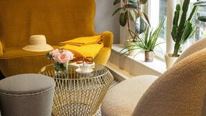 Uma sala de estar moderna com um sofá amarelo mostarda, uma poltrona amarela, um banquinho estampado e uma mesa de centro de vidro. Há duas luminárias fixadas na parede, uma janela grande com vista e uma planta verde alta.