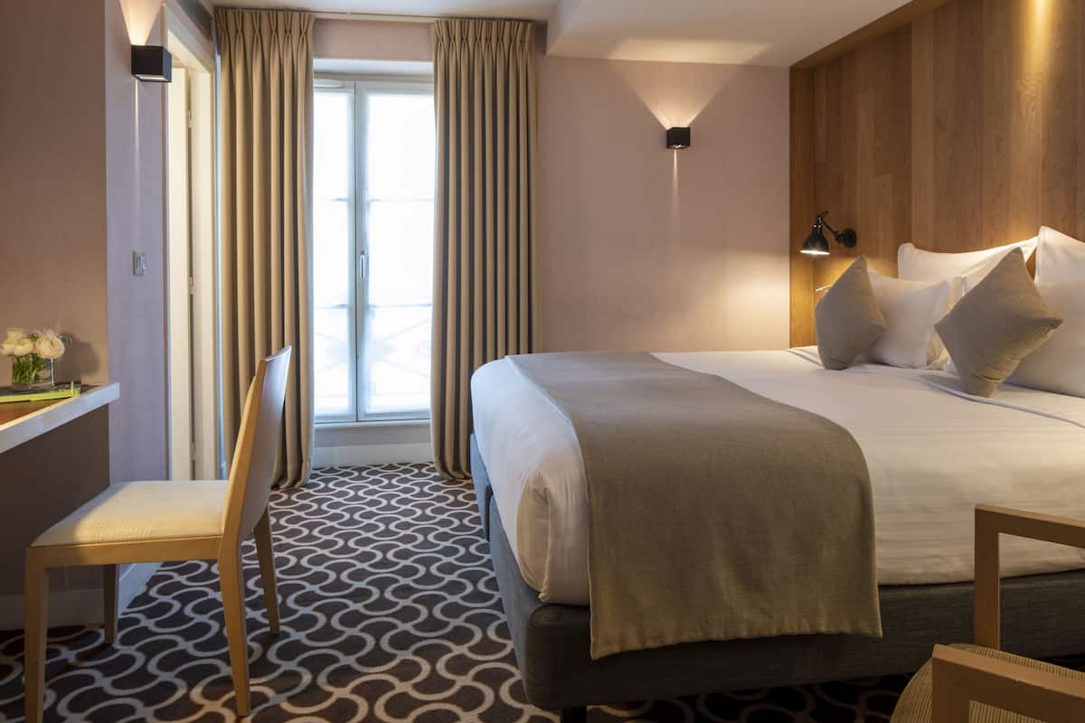 A hotel room with a bed, a chair, a desk with a lamp, and a window with curtains.