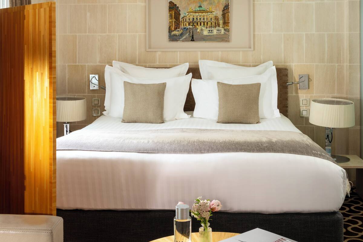 A hotel room with a large bed, two bedside lamps, a framed picture on the wall, a round wooden table with a book and a vase, and a large hanging light fixture.