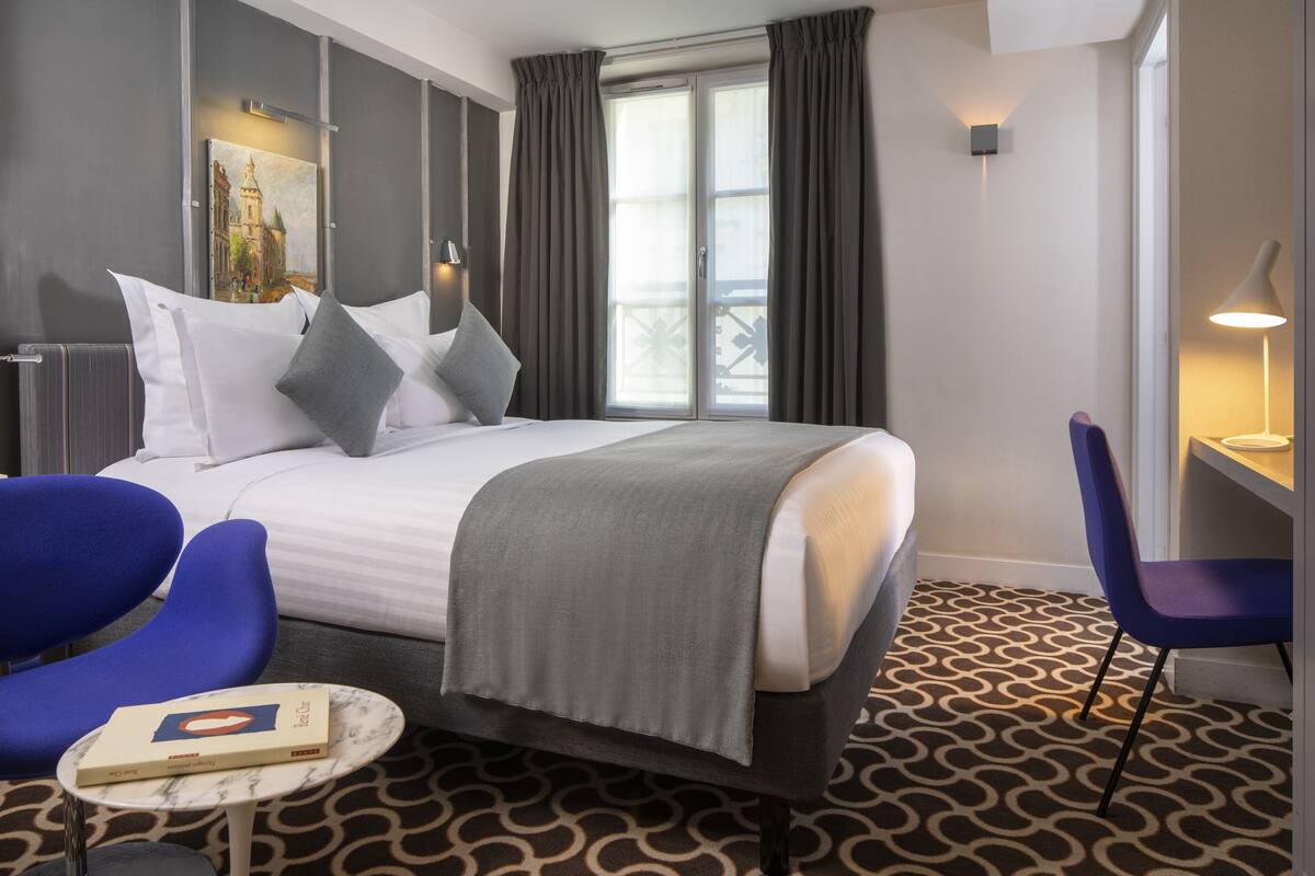A hotel room with a large bed, a small table with a book, a blue chair, and a window with curtains.