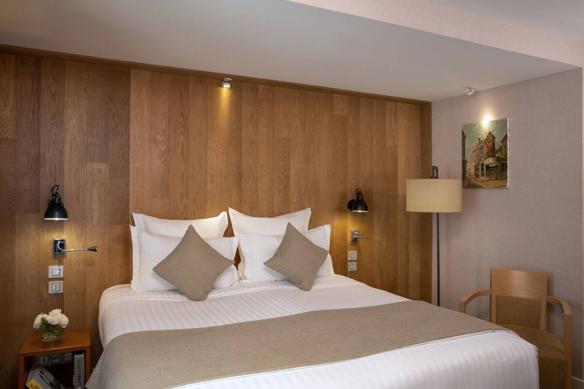 A hotel room with a wooden headboard, a bed with white linens, two bedside lamps, a nightstand with a vase of flowers, and a painting on the wall.