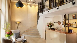 Um saguão de hotel com balcão de recepção, escada e área de estar com uma cadeira e uma mesa pequena.