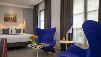 A hotel room with a bed, two blue armchairs, a small table with a magazine, and a painting on the wall.