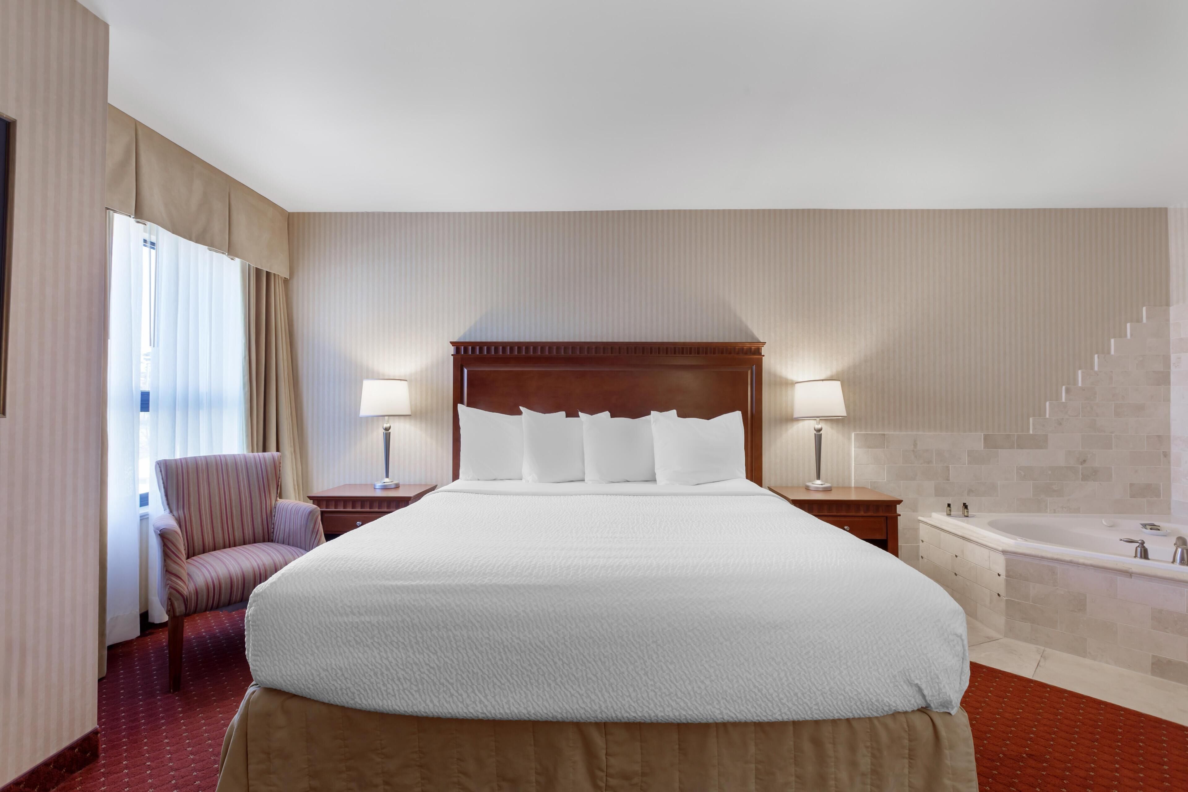 A hotel room with a large bed, a chair, a nightstand, a lamp, a bathroom with a bathtub, and a staircase.