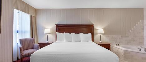 Quarto de hotel com cama grande, uma cadeira, mesa de cabeceira, abajur, banheiro com banheira e uma escada.