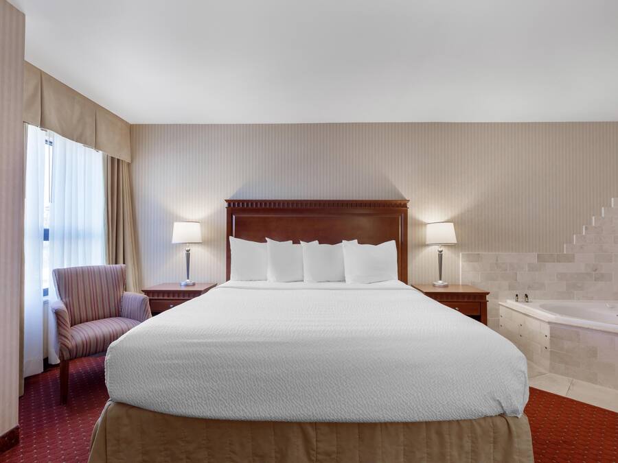 A hotel room with a large bed, a chair, a nightstand, a lamp, a bathroom with a bathtub, and a staircase.