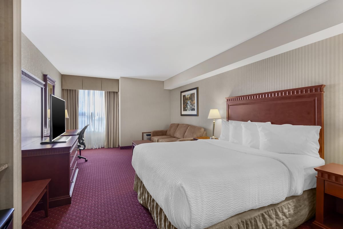 Ein Hotelzimmer mit einem großen Bett, einem Schreibtisch mit Computer, einem Stuhl, einer Lampe und einem gerahmten Bild an der Wand.