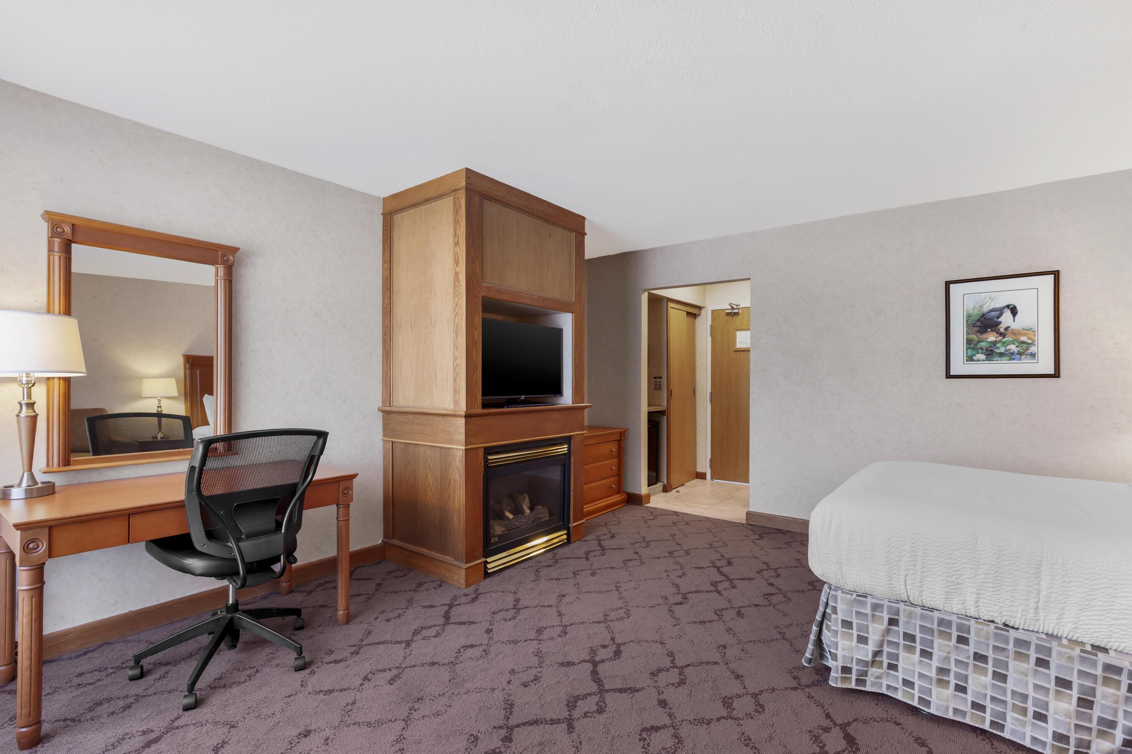 A hotel room with a wooden desk, a chair, a fireplace, a bed, a mirror, and a framed picture on the wall.