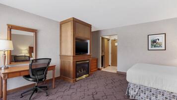 A hotel room with a wooden desk, a chair, a fireplace, a bed, a mirror, and a framed picture on the wall.