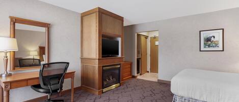 A hotel room with a wooden desk, a chair, a fireplace, a bed, a mirror, and a framed picture on the wall.