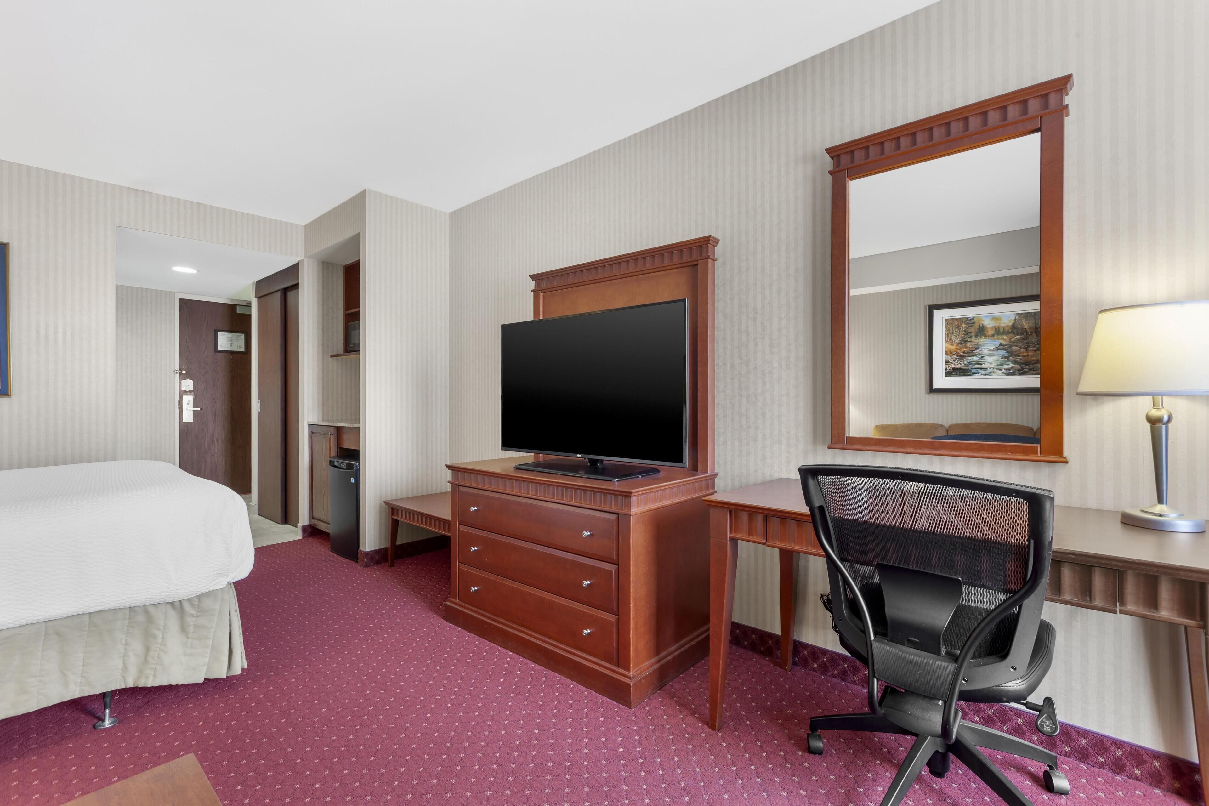 A hotel room with a bed, a desk with a computer, a television, a chair, a dresser, and a mirror.