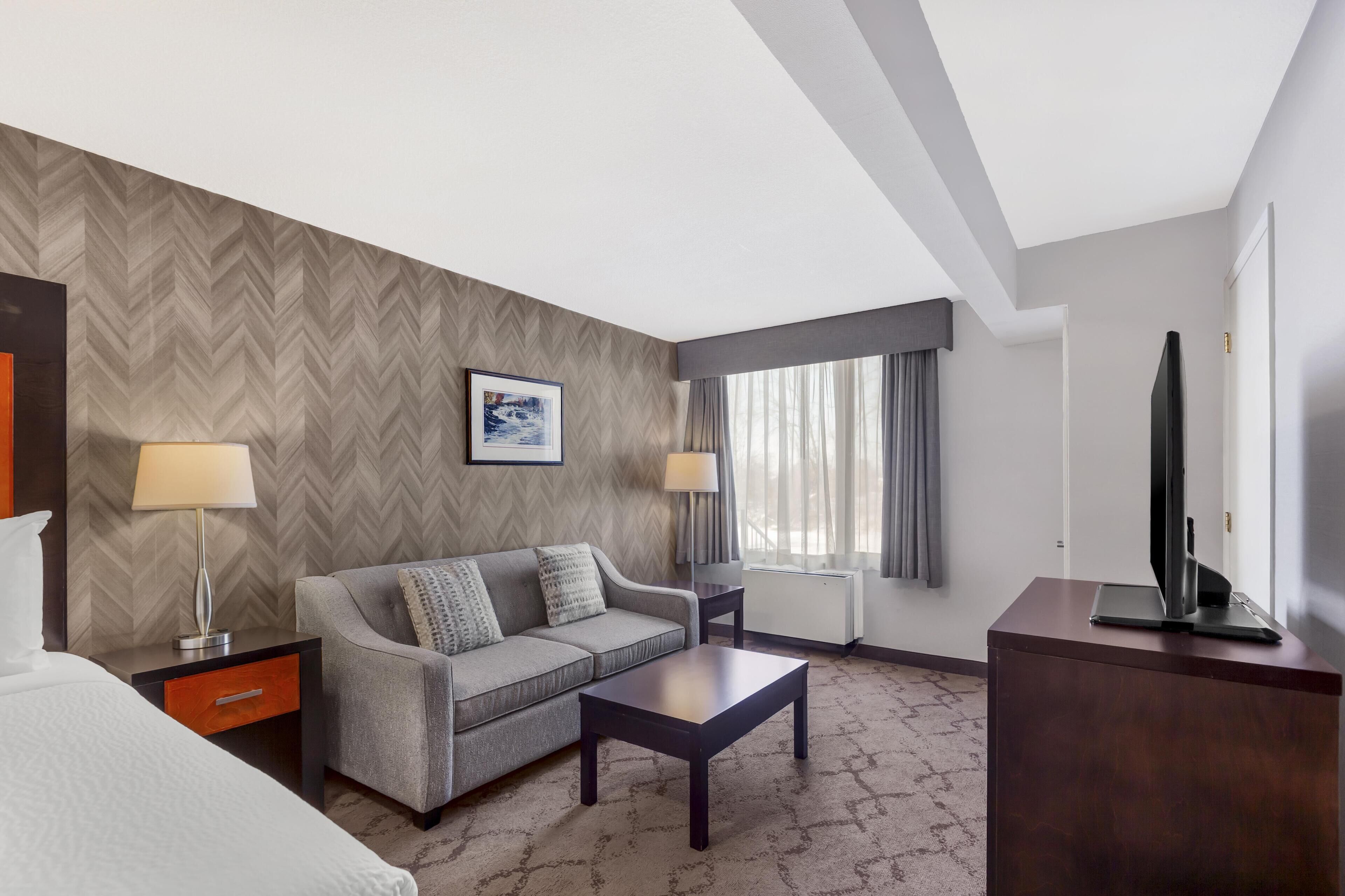 Ein Hotelzimmer mit Sofa, Couchtisch, Fernseher auf einem Schrank, Lampe, Bild an der Wand und einem Fenster mit Vorhängen.