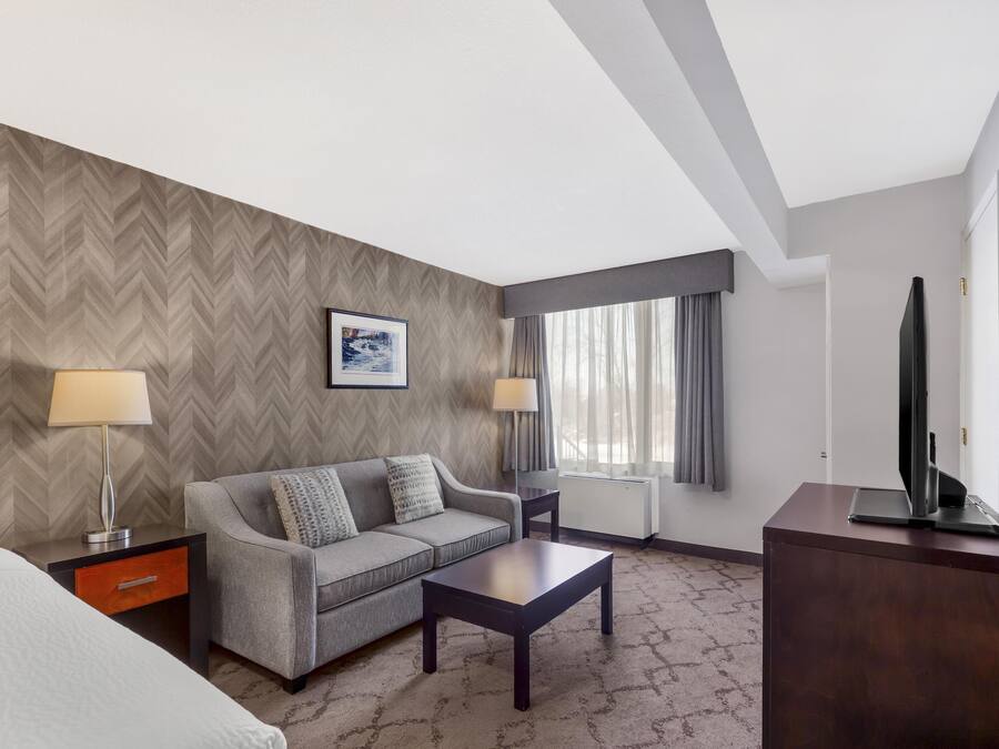 Ein Hotelzimmer mit Sofa, Couchtisch, Fernseher auf einem Schrank, Lampe, Bild an der Wand und einem Fenster mit Vorhängen.