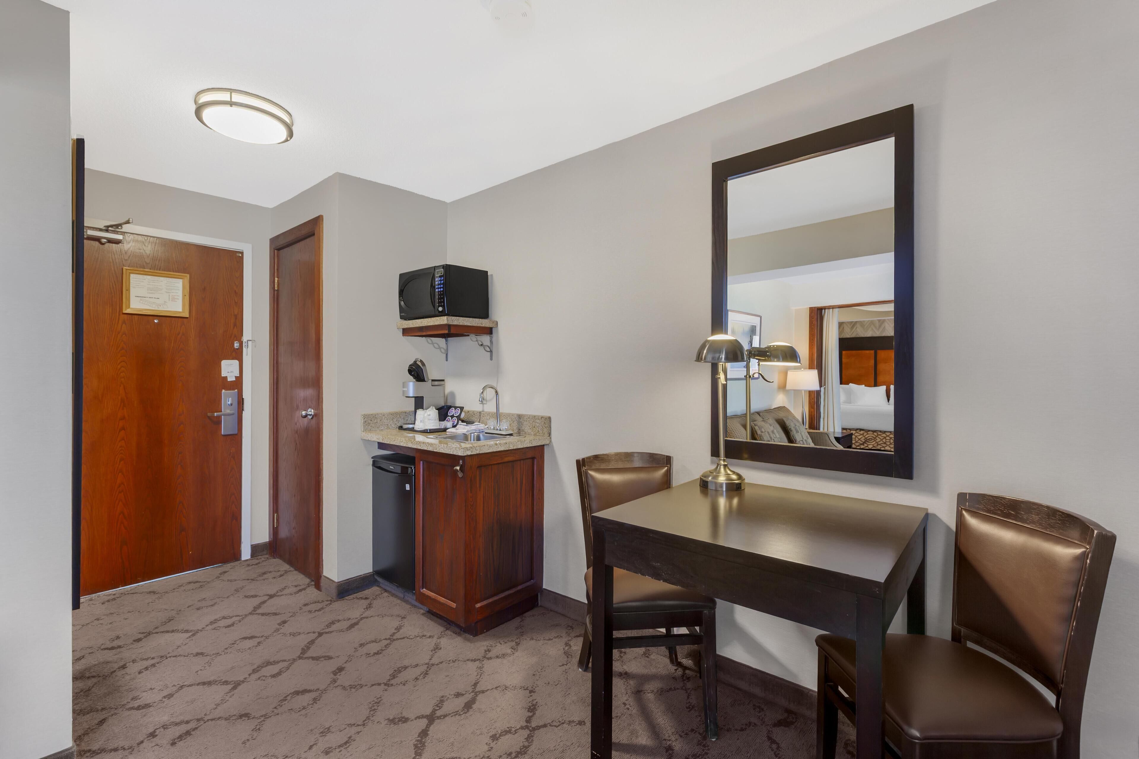 Une chambre d’hôtel avec une porte en bois, une kitchenette équipée d’un four à micro-ondes, d’un évier et d’un petit placard. On y trouve une table à manger avec deux chaises et un grand miroir accroché au mur.
