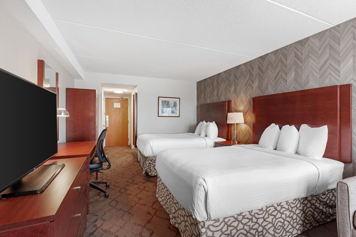 Ein Hotelzimmer mit zwei Betten, einem Schreibtisch, einem Flachbildfernseher, einem Sessel und einem gemusterten Teppich.