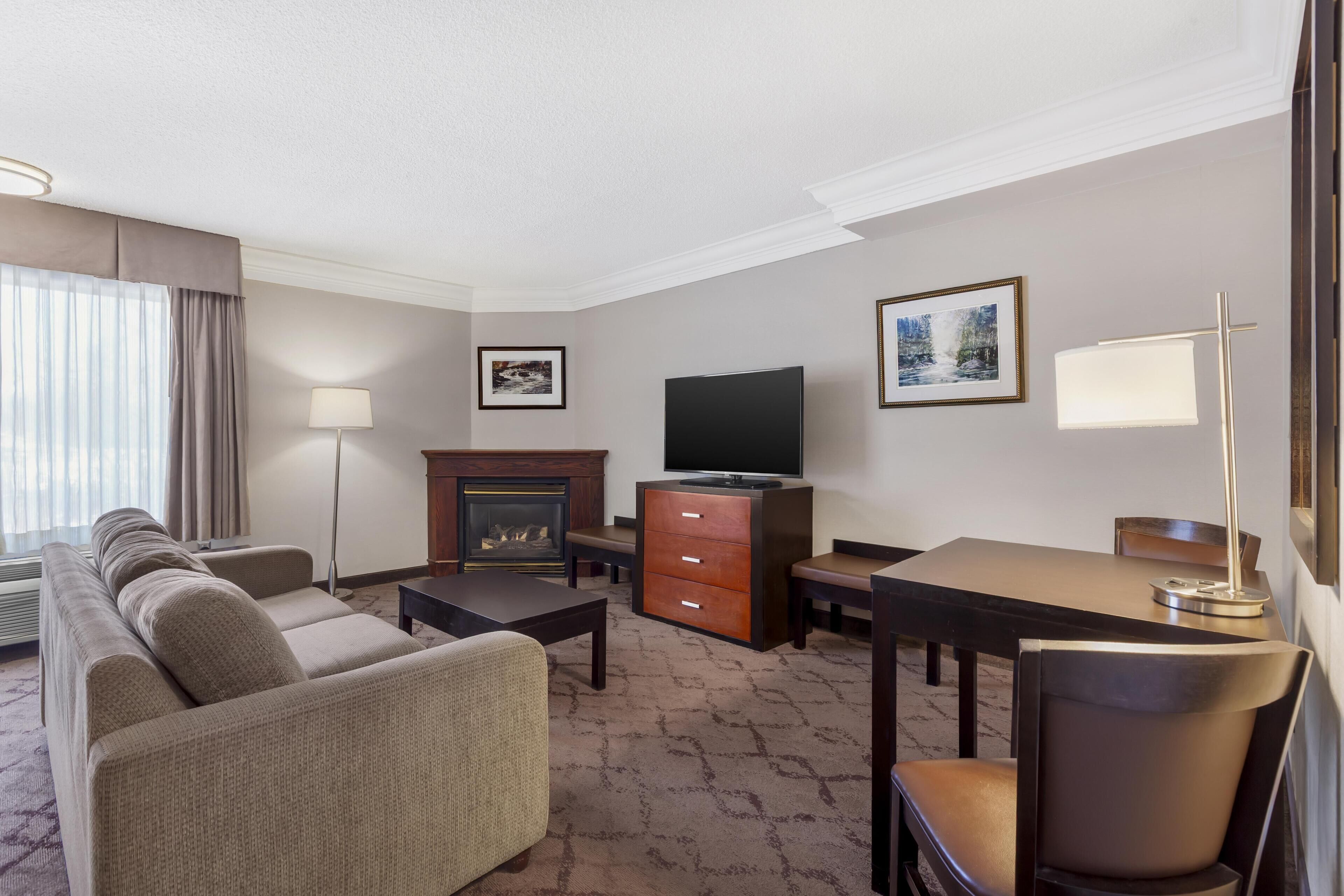A hotel room with a sofa set, a flat-screen TV, a fireplace, a desk, and a lamp.