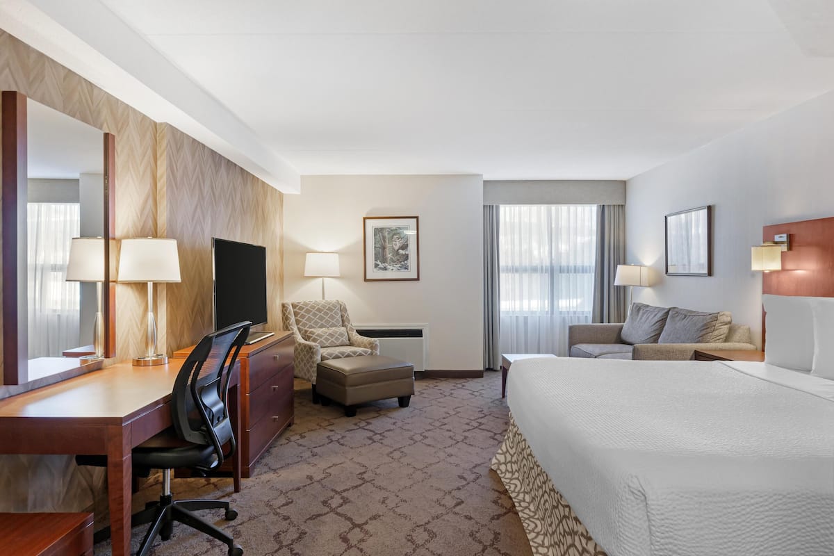 Ein Hotelzimmer mit einem großen Bett, einem Schreibtisch mit Stuhl, einer Couch, einem Sessel, einem Fernseher und einem Fenster mit Vorhängen.